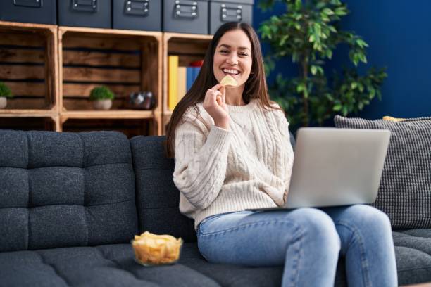 mujer joven usando teléfono inteligente y comiendo papas fritas en casa - potatoe chips fotografías e imágenes de stock