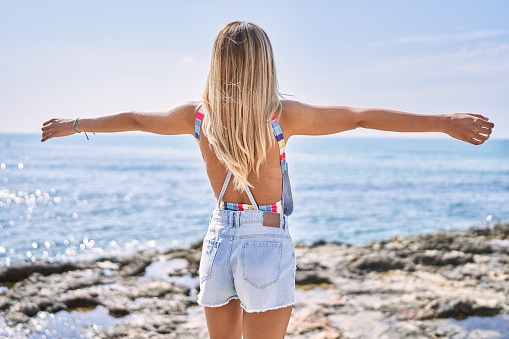 Young blonde girl on back view breathing with arms open standing at the beach.