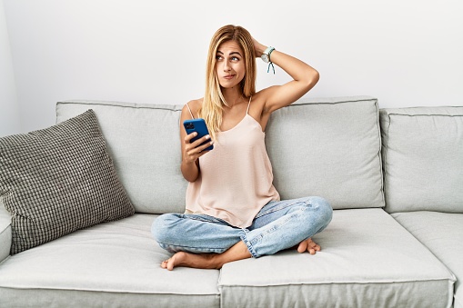 Blonde beautiful young woman sitting on the sofa at home using smartphone confuse and wondering about question. uncertain with doubt, thinking with hand on head. pensive concept.