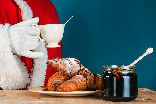 The hand of Santa Claus in traditional costume holds a porcelain white cup. Close-up pot with honey, croissants with sugar powder on the table. Dark turquoise, background with copy space.