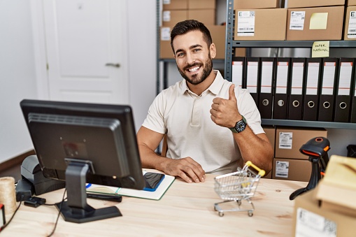 Handsome hispanic man working at small business commerce doing happy thumbs up gesture with hand. approving expression looking at the camera showing success.