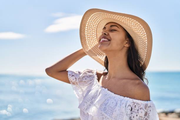 joven turista afroamericana sonriendo confiada usando sombrero de verano a la orilla del mar - women summer hat beach fotografías e imágenes de stock