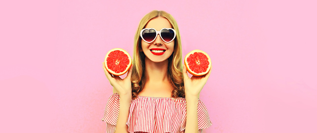 Summer portrait of happy smiling young woman with slice of juicy grapefruit wearing heart shaped sunglasses on pink background