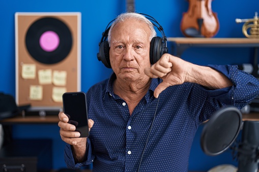 Senior man with grey hair showing smartphone screen at music studio with angry face, negative sign showing dislike with thumbs down, rejection concept