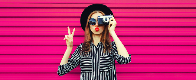 Portrait of stylish modern young woman photographer with film camera blowing her lips on pink background