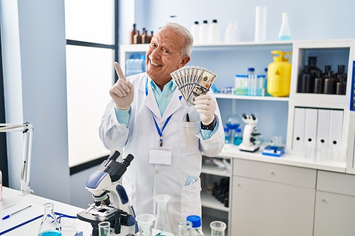 Senior scientist with grey hair working at laboratory holding dollars smiling happy pointing with hand and finger to the side