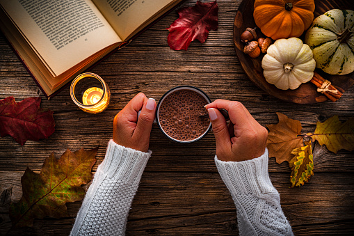 Autumn backgrounds:  woman's hands holding hot chocolate mug shot from above on rustic wooden table. Dry leaves, mini pumpkins and an open book complete the composition. High resolution 42Mp studio digital capture taken with SONY A7rII and Zeiss Batis 40mm F2.0 CF lens