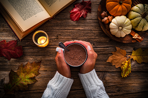 Autumn backgrounds:  woman's hands holding hot chocolate mug shot from above on rustic wooden table. Dry leaves, mini pumpkins and an open book complete the composition. High resolution 42Mp studio digital capture taken with SONY A7rII and Zeiss Batis 40mm F2.0 CF lens