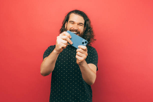 Photo of joyful young bearded hispter man with long hair playing online games at his smartphone over pink background. stock photo