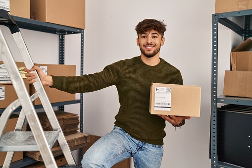 Young man ecommerce business worker holding package standing on ladder at office