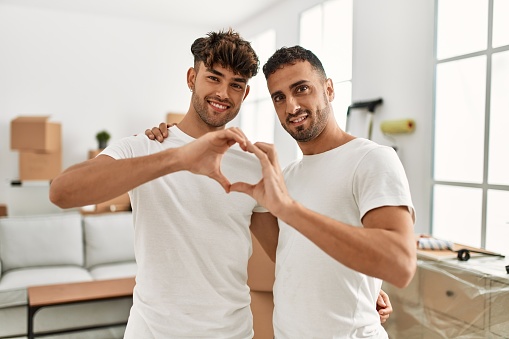 Two hispanic men couple hugging each other doing heart shape with hands at new home