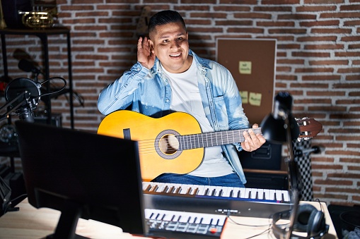 Hispanic young man playing classic guitar at music studio smiling with hand over ear listening an hearing to rumor or gossip. deafness concept.