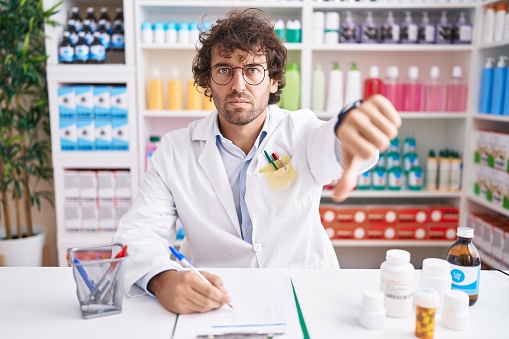 Hispanic young man working at pharmacy drugstore looking unhappy and angry showing rejection and negative with thumbs down gesture. bad expression.