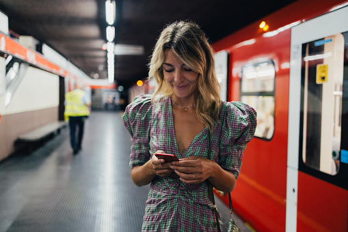 A smiling Caucasian influencer watching something on her smartphone while waiting for the train.