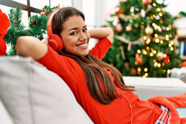 jeune femme latine écoutant de la musique assise près du sapin de noël à la maison - christmas tree audio photos et images de collection