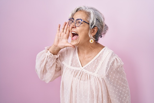 Middle age woman with grey hair standing over pink background shouting and screaming loud to side with hand on mouth. communication concept.