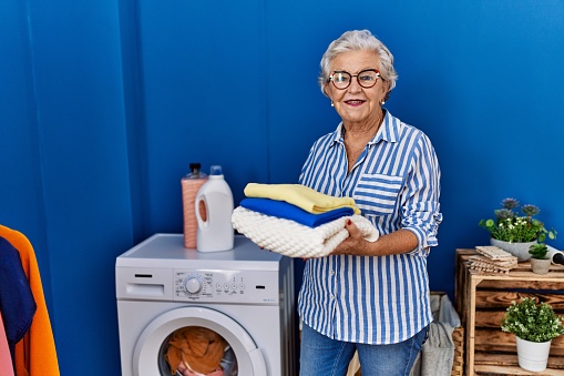 Senior grey-haired woman smiling confident holding folded clothes at laundry room