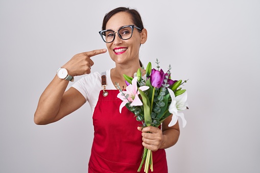 Middle age brunette woman wearing apron working at florist shop holding bouquet smiling cheerful showing and pointing with fingers teeth and mouth. dental health concept.