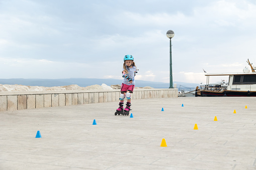 Little girl is rollerblading along the embankment. The child rolls between the colored chips. The child uses a helmet and protection for elbows and knees.