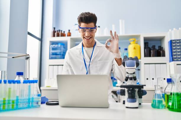 giovane uomo ispanico che lavora al laboratorio di scienziati facendo videochiamata facendo ok segno con le dita, sorridente amichevole gesticolando simbolo eccellente - spanish and portuguese ethnicity immagine foto e immagini stock