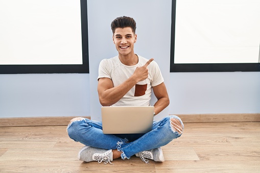 Young hispanic man using laptop at home cheerful with a smile on face pointing with hand and finger up to the side with happy and natural expression