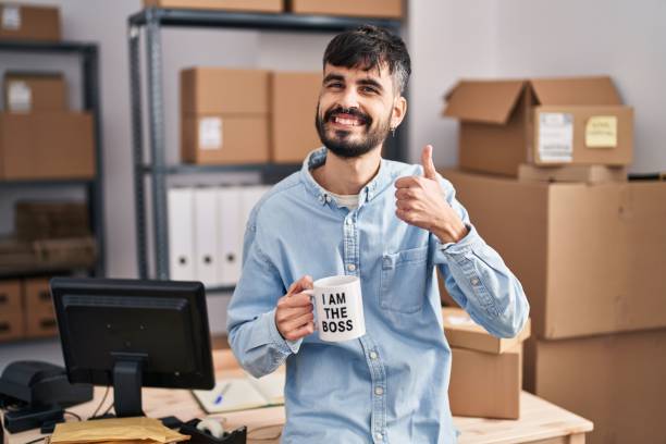 istock Young hispanic man with beard working at small business ecommerce drinking from boss cup smiling happy and positive, thumb up doing excellent and approval sign 1436422614