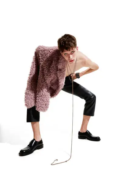 Photo of Portrait of expressive young man posing shirtless in leather pants and pink furry coat over white background