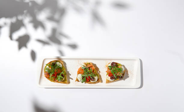 istock Aesthetic composition with salmon bruschetta on white background with shadows from flowers. Italian bruschetta with salmon, avocado, cheese and olives on fine dining in summer. Elegant menu concept. 1436425604