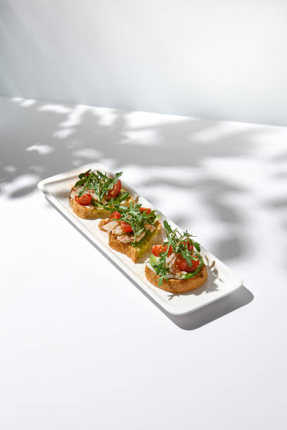 istock Aesthetic composition with shrimp bruschetta on white background with shadows from flowers. Italian bruschetta with shrimp, tomatoes and arugula on fine dining in summer. Elegant menu concept. 1436425617