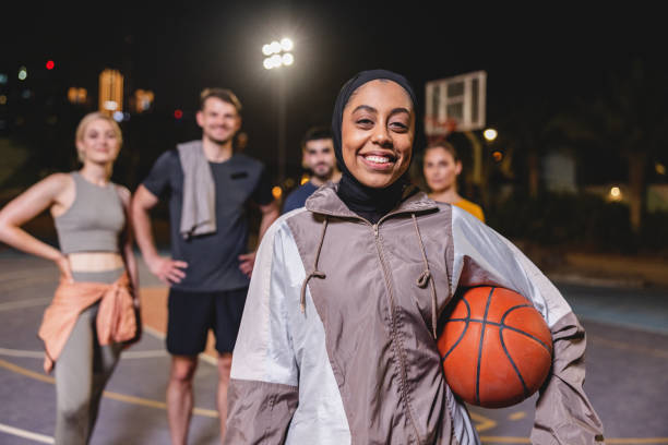 Portrait Of Happy Middle Eastern Adult Female Holding Basketball While Her Team Is Standing Behind Her On Basketball Court stock photo