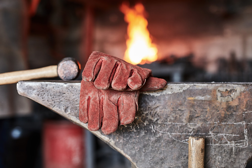 Gloves, hammer and anvil in the workshop of an artisan for craftsmanship with a fire in the background. Industrial, manufacturing and production with equipment in a forge of foundry for creativity