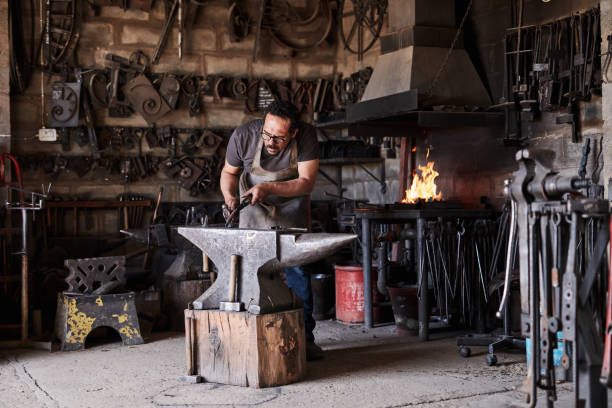 istock Blacksmith man in workshop, metal tools with fire and industrial building. Career in manufacturing steel products, handmade iron equipment and forge with hammer in flame and construction in London 1436428971