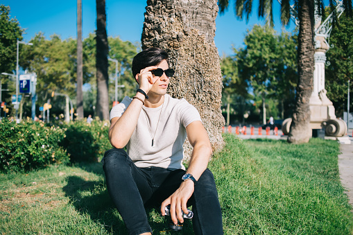 Confident ethnic male wearing casual clothes and stylish sunglasses sitting with vintage photo camera on green lawn in park while relaxing during city stroll in Barcelona and looking away
