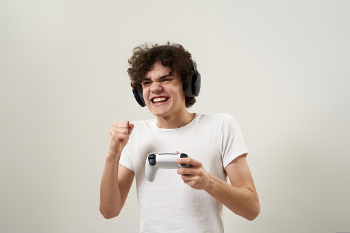 Excited caucasian teenage boy celebrate his win in video game with joystick. Curly guy of zoomer generation wear t-shirt and headphones. Youngster lifestyle. White background. Studio shoot. Copy space