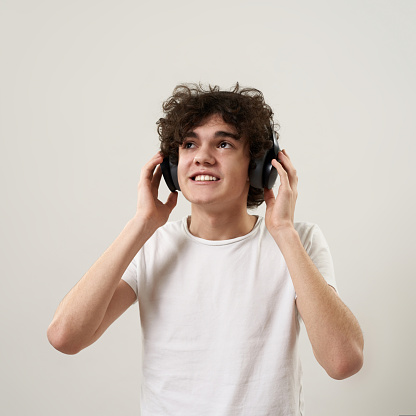 Caucasian teenage boy listening music in headphones. Curly guy of zoomer generation wearing t-shirt. Concept of modern youngster lifestyle. Isolated on white background. Studio shoot. Copy space