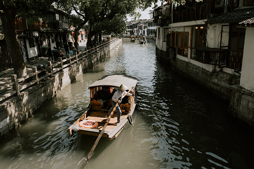 A  close shot of the landscape or cityscape of Chinese fishermen paddling on their boats on a river or canal under the tree between the short Chinese traditional ancient houses or historical architectures or old residential buildings with balcony in an old fishing village, showing the lifestyle, culture and history of China, during day time under the sun.