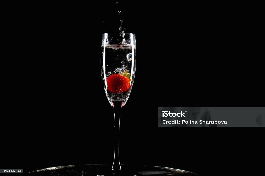 red strawberry falls into a glass with splashes on a black background red strawberry falls into a glass of water with splashes on a black background Champagne Stock Photo
