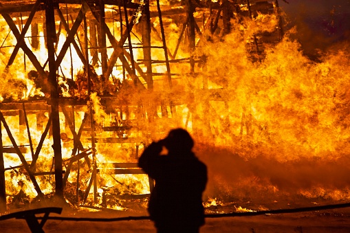 The black silhouette of a photographer's man against the backdrop of a huge burning flame. The dark figure of a man against the backdrop of a fire blast at night. A lonely man in front of raging fire. The end of times background. It is the end of the world.