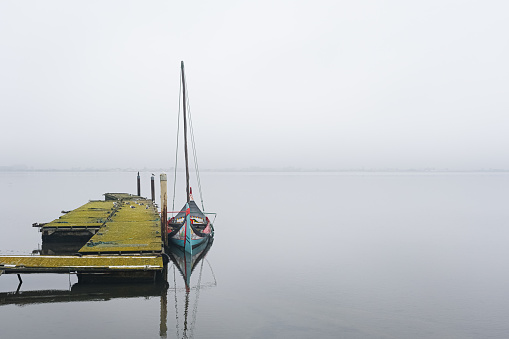 An old wooden pier and a brightly painted fishing boat next to it on the background of a foggy Atlantic ocean bay.