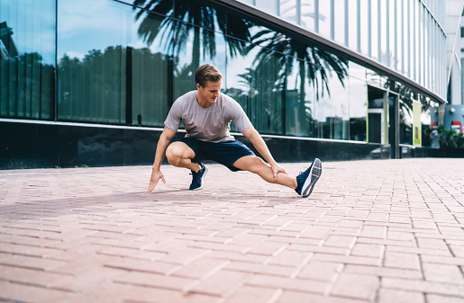 Strong caucasian male jogger making warming exercises stretching lower muscles on street, sportsman in active wear squatting on training keeping healthy lifestyle and goals for vitality and wellness