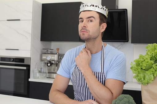 A majestic looking man is wearing a crown in the kitchen. He has some groceries in a paper bag and uses an apron.