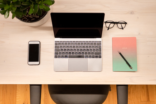 High angle view of a work desk with a smart phone, laptop computer, notepad and glasses.