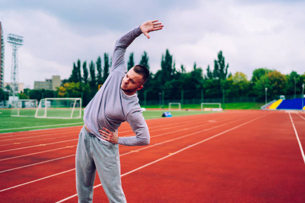 atleta maschio caucasico serio in abbigliamento sportivo che fa esercizi di lavoro prima di iniziare a correre allo stadio, forte sportivo degli anni '20 in tuta grigia che si allena al mattino mantenendo uno stile di vita sano - determination 20s adult attractive male foto e immagini stock