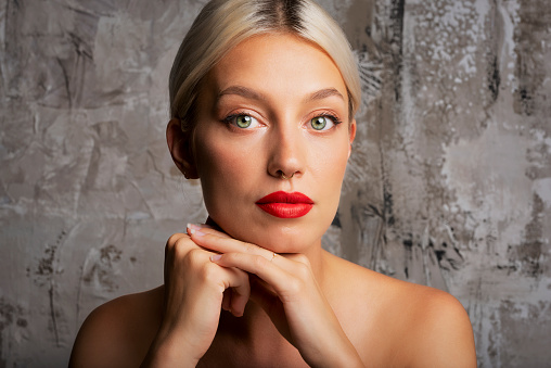 Headshot of gorgeous young woman with blond hair wearing red lipstick and nose piercing while standing at isolated grey background. Copy space.