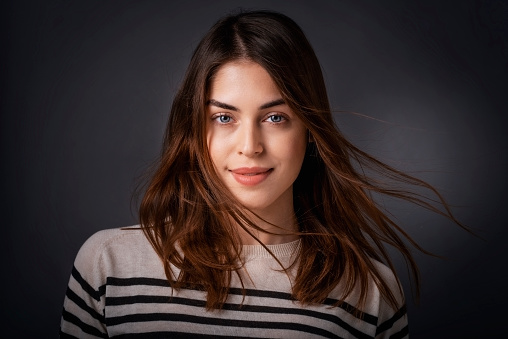 Studio portrait of beautiful young woman looking at camera and smiling while standing at isolated dark background.