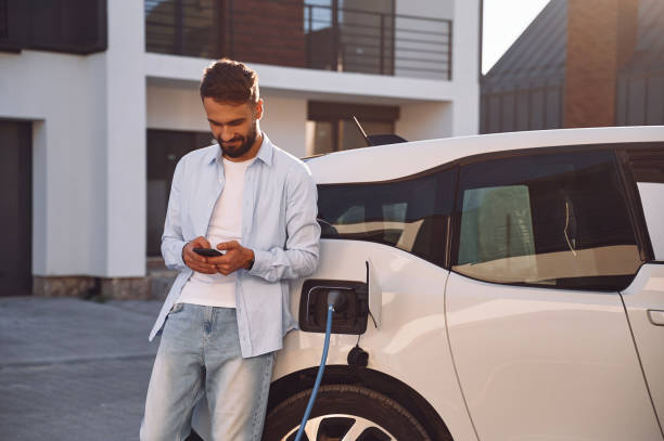 Charging the vehicle near the house. Holding smartphone. Young stylish man is with electric car at daytime stock photo
