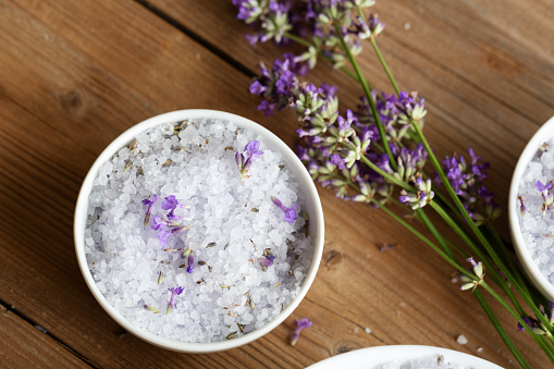 The relaxing power oft he lavender plant is used to add scent to sea salt in white bowls on wooden background, the salt containts dried lavender flowers and is decorated with a bunch of fresh lavender, seen from above