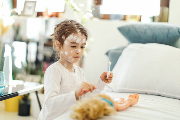 Girl has a chickenpox and spending time at home in quarantine. She is playing and applying to her doll same lotion that she has on her face