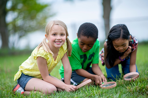 A small group of three school aged children kneel down in the grass together with magnifying glasses as they explore during day camp.  They are each dressed casually and are focused on finding bugs in the grass.