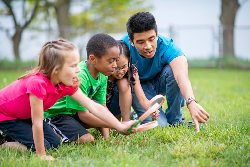 A male day camp counselor bends down in the grass with a small group of school aged children as they explore together with magnifying glasses.  They are each dressed casually and are wearing colorful t-shirts as they focus on their task.